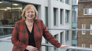 Christina McAnea leans against a railing in UNISON centre, wearing a red patterned jacket